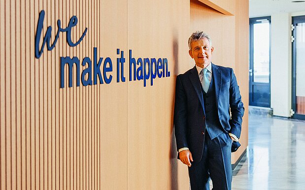 Wolfgang Litzlbauer steht in einem modernen Bürogebäude vor einer Holzvertäfelung mit dem Schriftzug „we make it happen“.