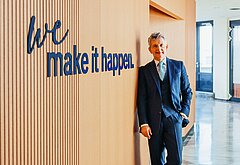 Wolfgang Litzlbauer steht in einem modernen Bürogebäude vor einer Holzvertäfelung mit dem Schriftzug „we make it happen“.