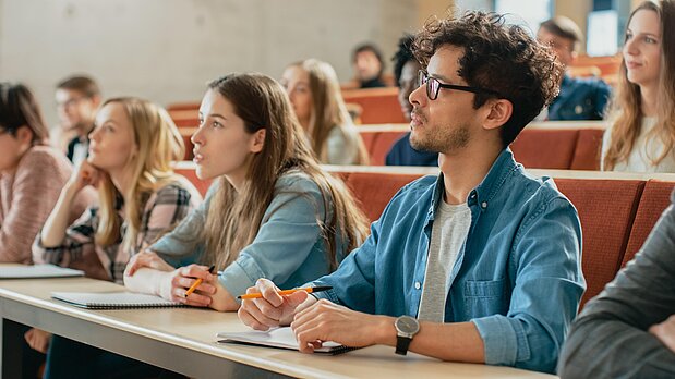Mehrere Studierende sitzen in einem Hörsaal und hören einem Dozenten zu. 