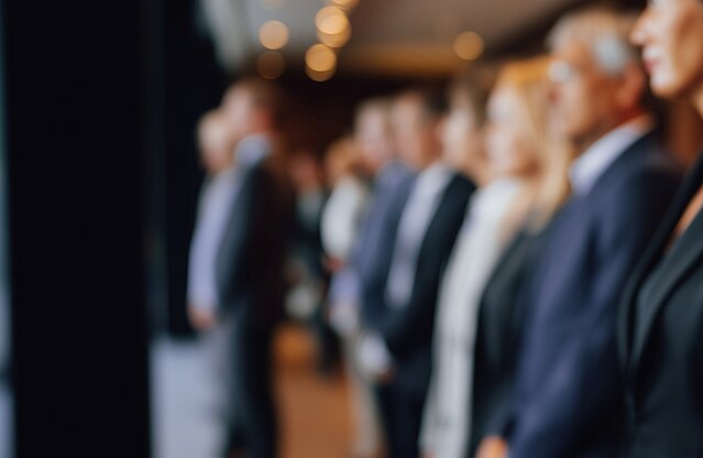Reihe elegant gekleideter Personen im Profil, Blick nach vorne, unscharfer Konferenzraum mit warmen Deckenlichtern.