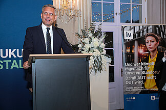 Wirtschaftsminister Wolfgang Hattmannsdorfer spricht am Rednerpult bei der Eröffnung des „Tags der NÖ Industrie“ in Laxenburg; im Hintergrund ein Plakat der IV-NÖ-Kampagne „Damit AUT nicht OUT wird“.